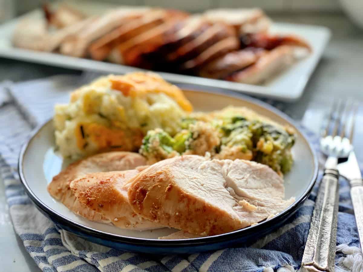 Three slices of turkey breast on a plate filled with side dishes with silverware and napkin on the side.