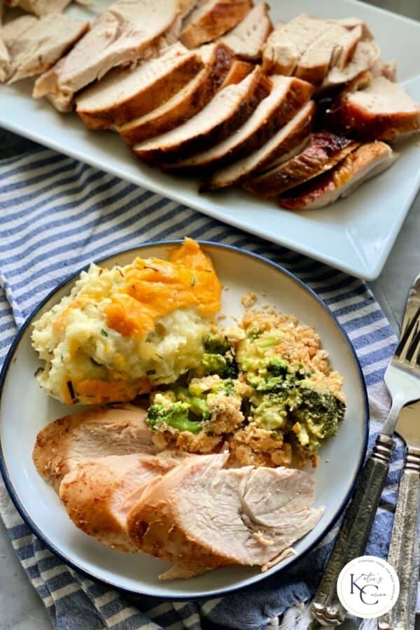 Top view of a white plate filled with turkey breast slices with mashed potatoes and broccoli casserole with a platter filled with sliced turkey breast.