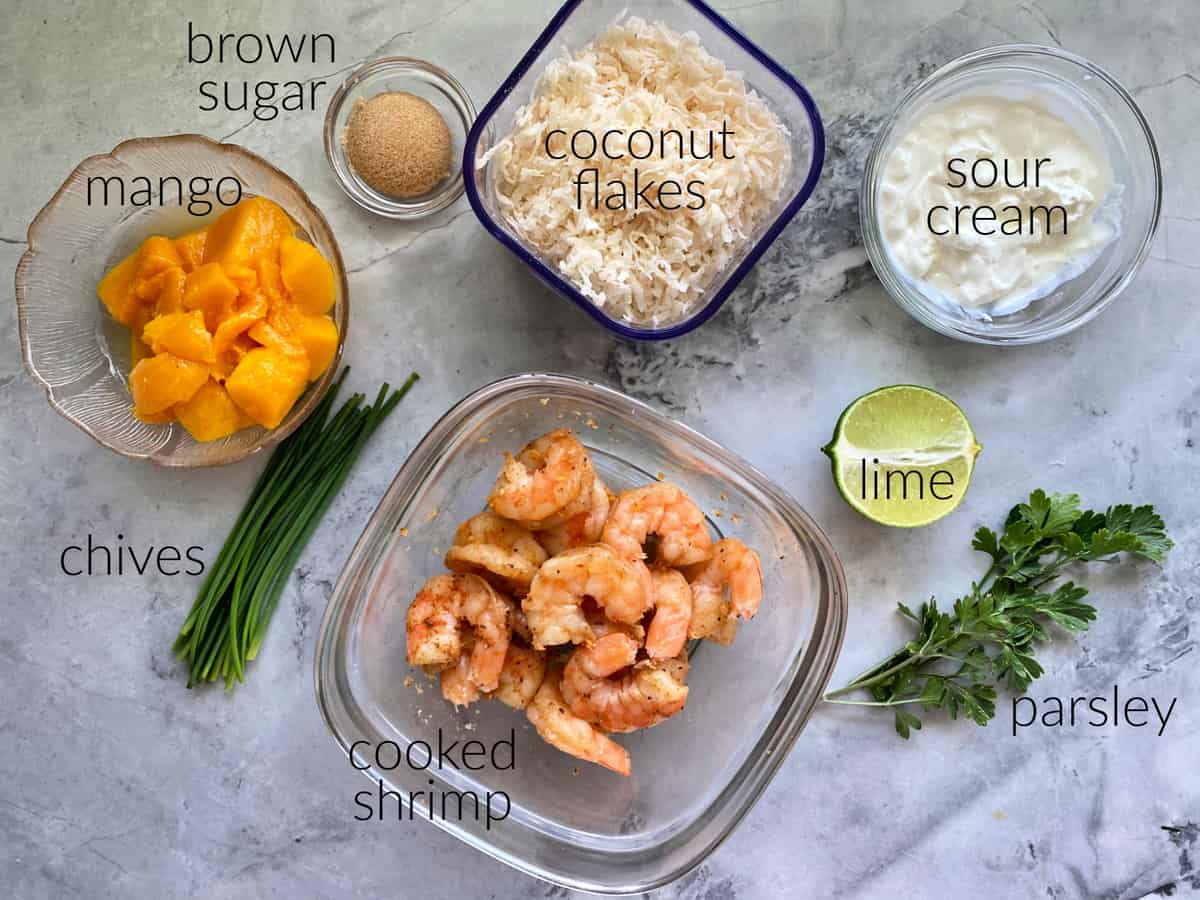 Ingredients on countertop: mango, brown sugar, chives, shrimp, coconut flakes, sour cream, lime, parsley.