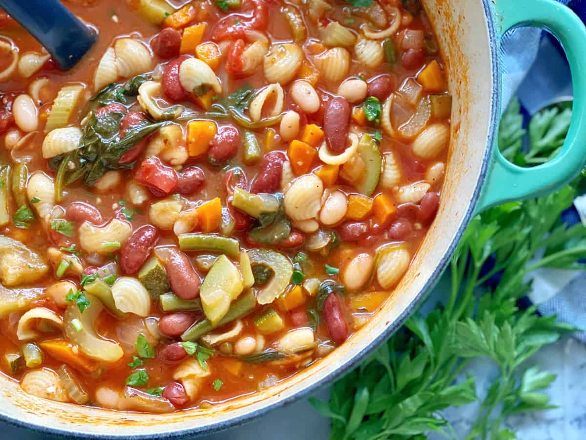 Top view of a green pot filled with minestrone soup with mini shell pasta with parsley next to it.