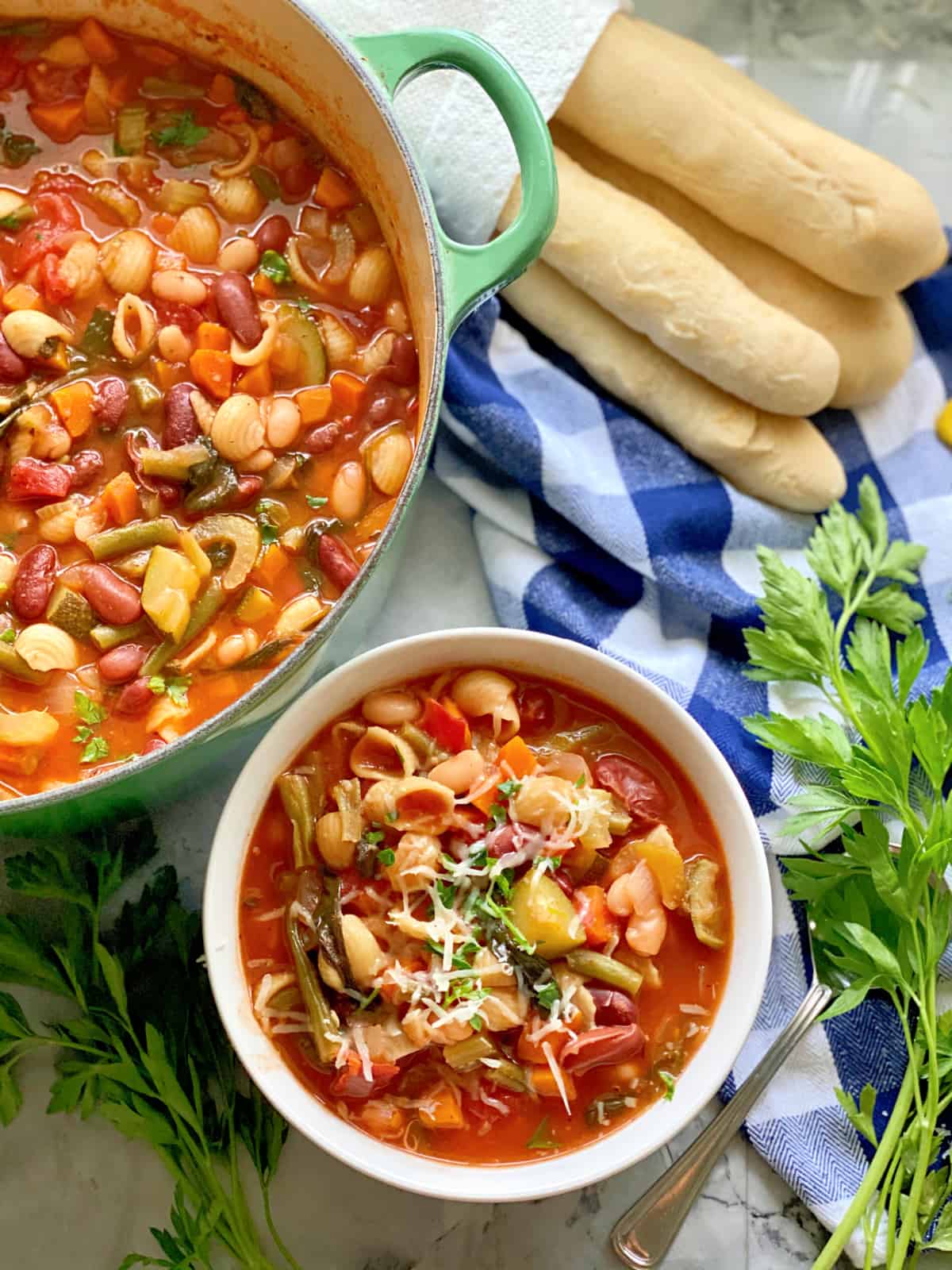 Top view of a large pot filled with minestrone soup with a bowl filled with soup and breasticks next to it.