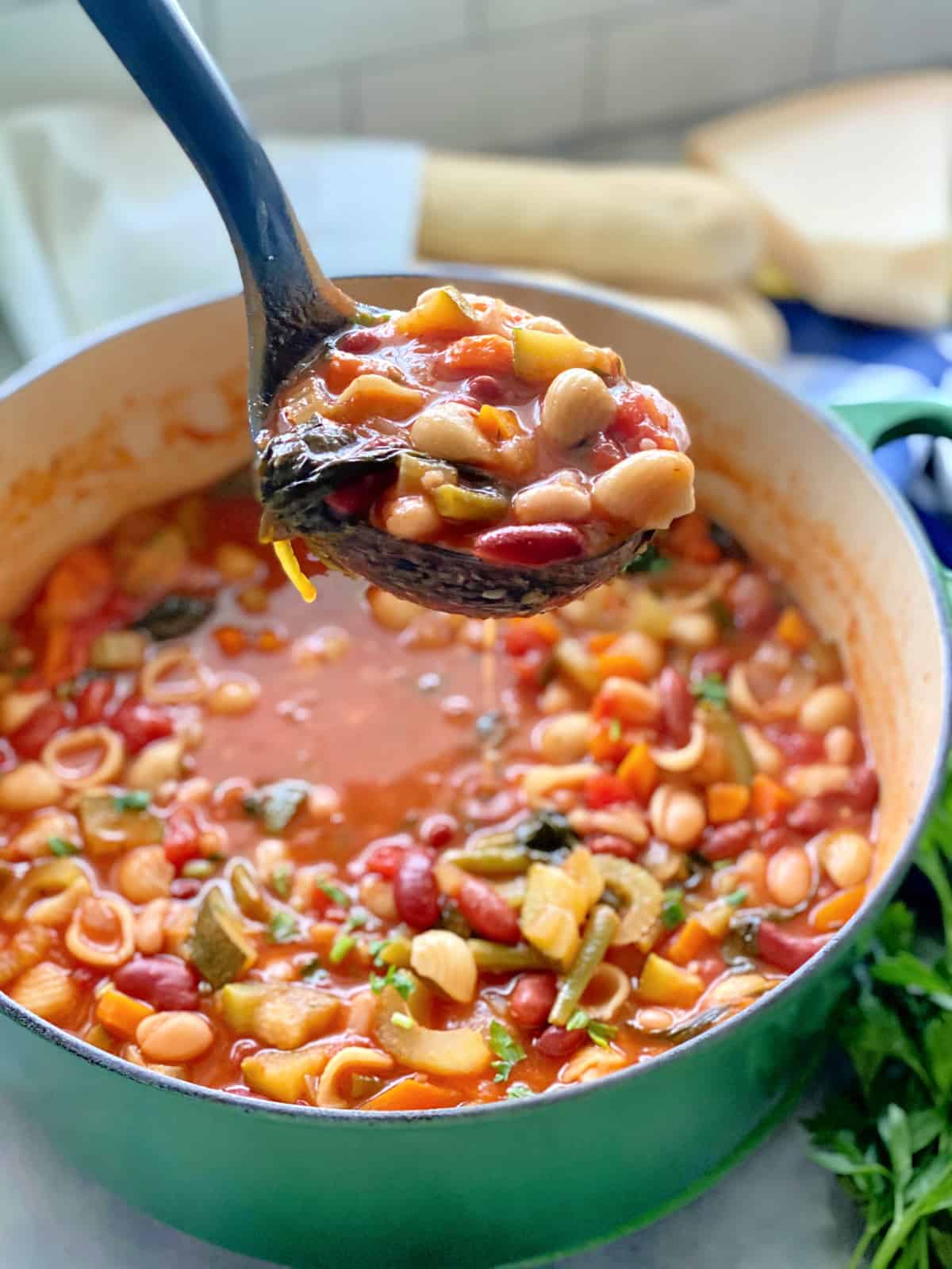 Green pot filled with minestrone soup and a black ladle filled with minestrone over the pot.