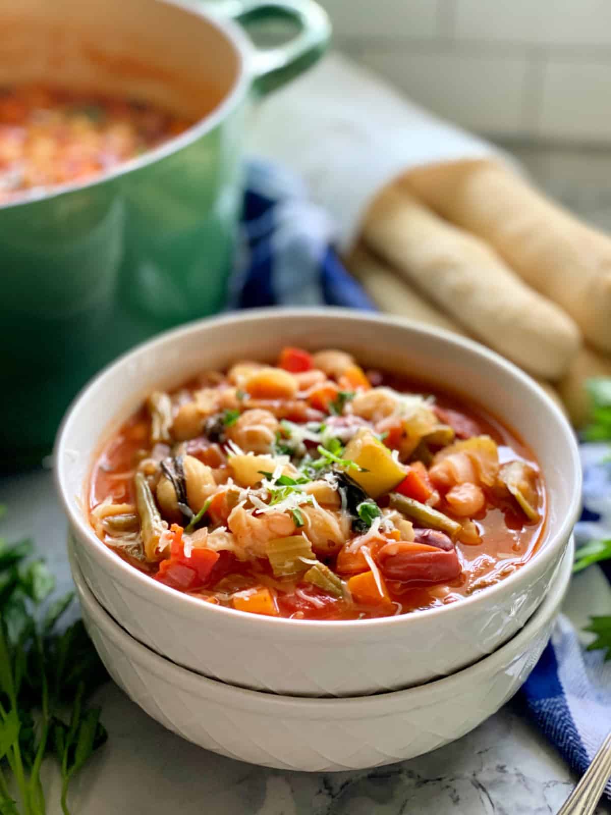 Two white bowls filled with minestrone soup with green pot and breadsticks in the background.