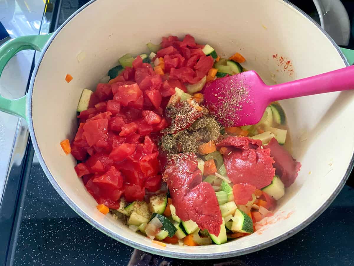 White pot filled with spices, tomato pasta, diced tomatoes, and vegetables with a pink spatula.