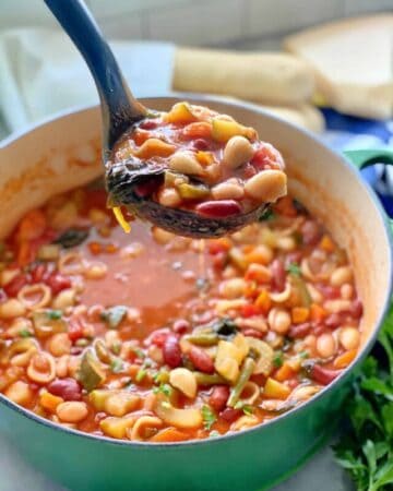 Green pot filled with minestrone soup and a black ladle filled with minestrone over the pot.