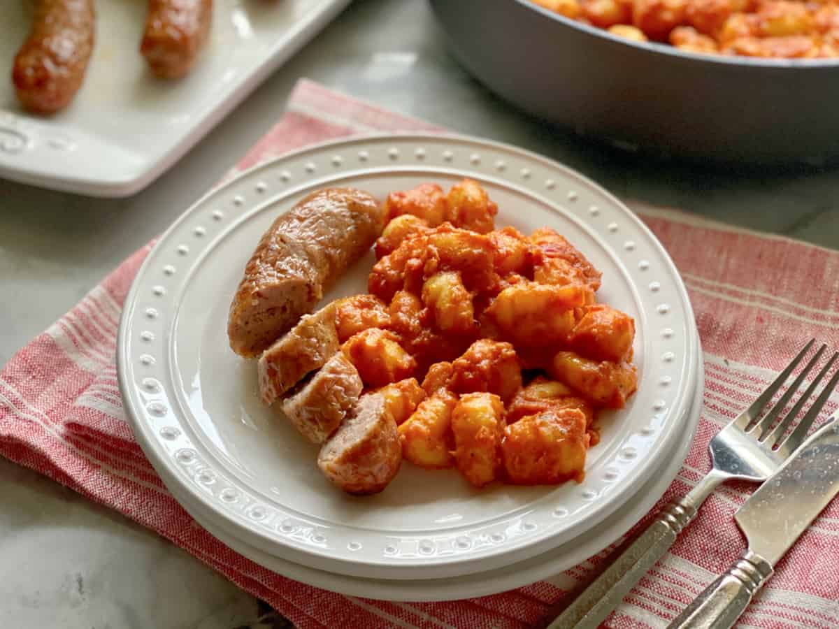 Two white plates filled with gnocchi in red sauce with sliced sausage with a fork and knife next to it.