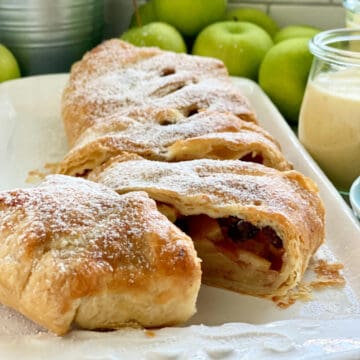 White platter with a puff pastry Apple Strudel sliced with apples in background.