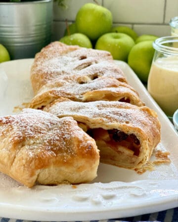 White platter with a puff pastry Apple Strudel sliced with apples in background.