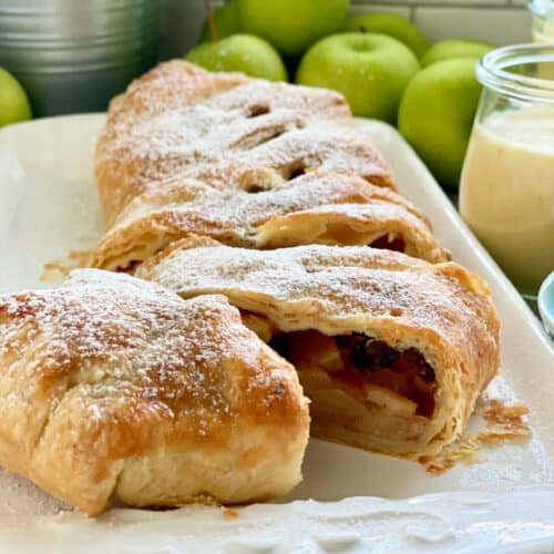 White platter with a puff pastry Apple Strudel sliced with apples in background.