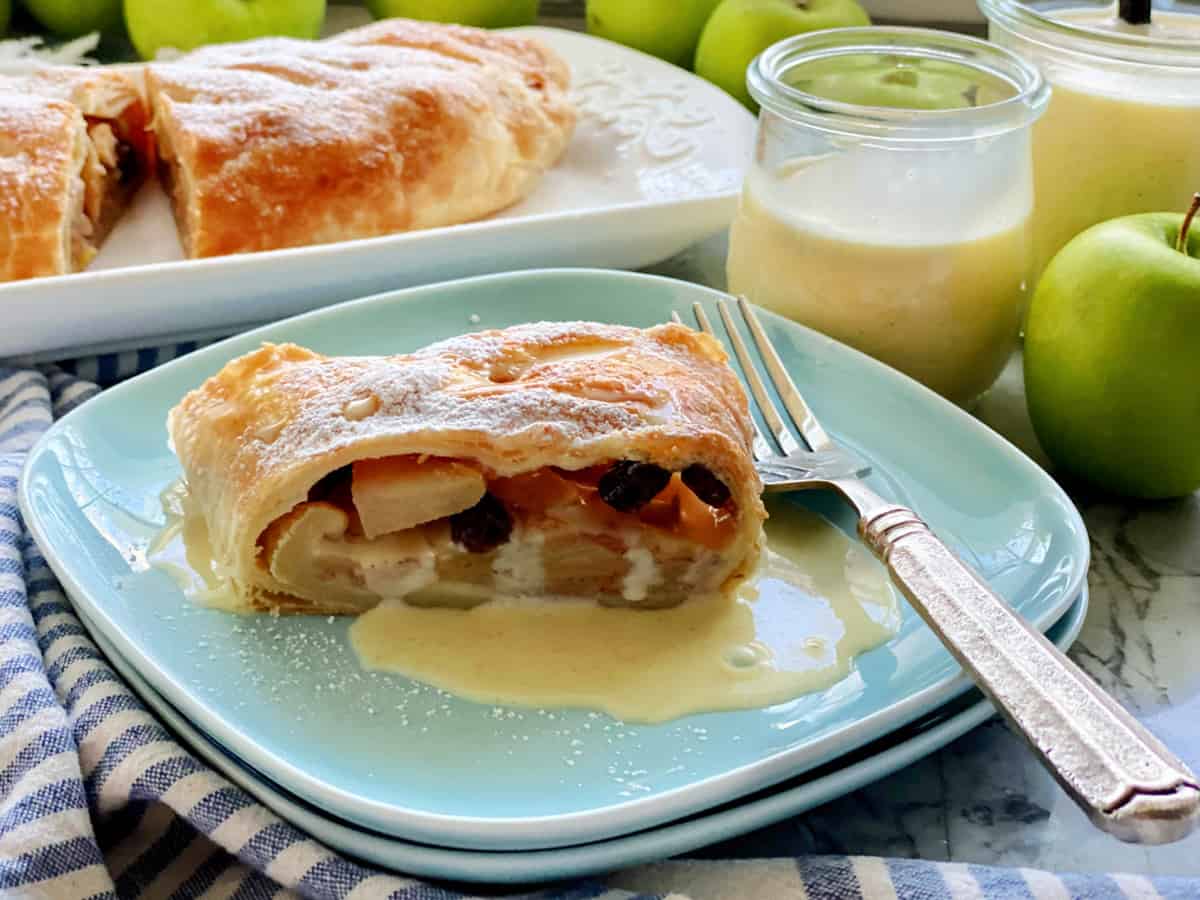 Two blue square plates with a slice of Apple Strudel with vanilla sauce and a fork on the plate.