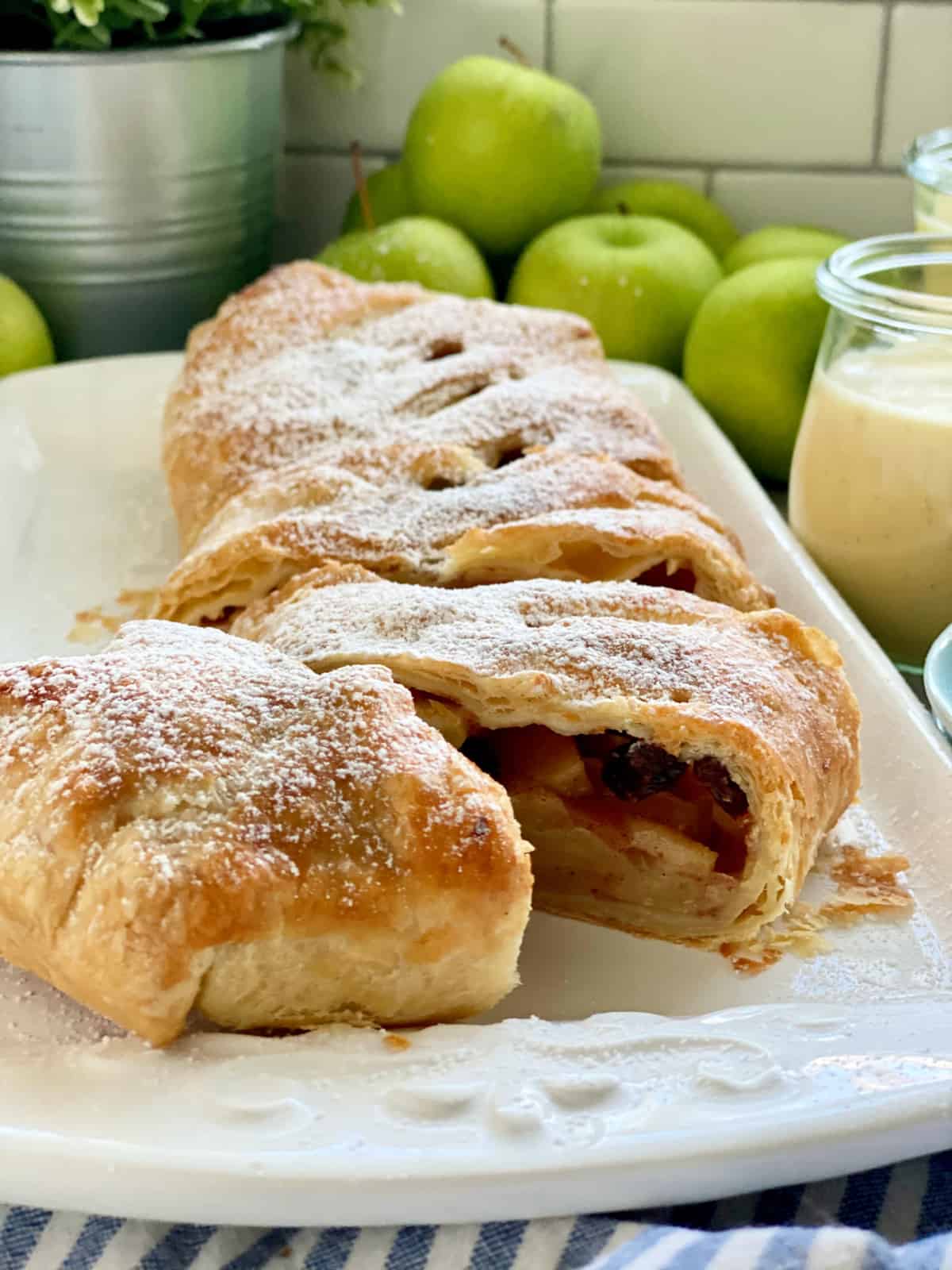 White platter with a puff pastry Apple Strudel sliced with apples in background.