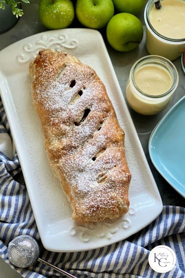 Top view of a white platter with a powder sugar dusted Apple Strudel with logo on right corner.