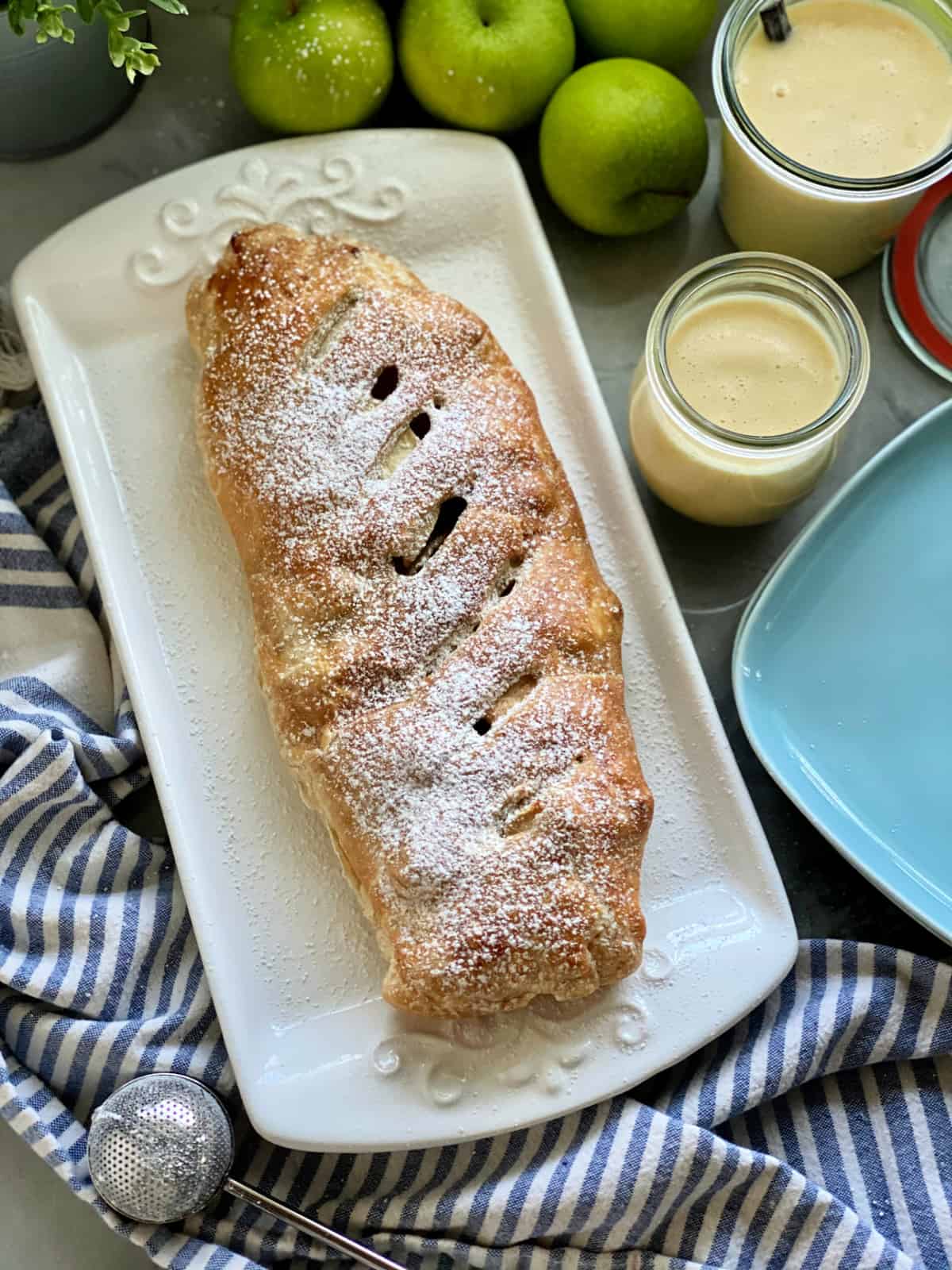 Top view of a white platter filled with an Apple Strudel with powder sugar, apples, and vanilla sauce on the side.