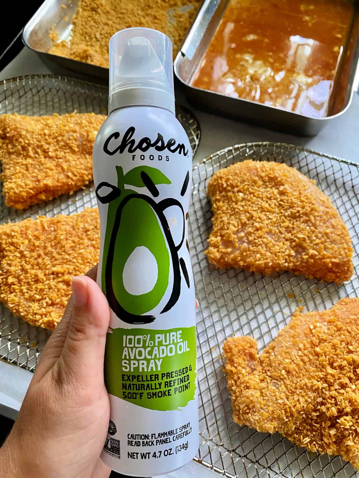 Female hand holding a can of avocado oil spray above trays of raw pork chops. 