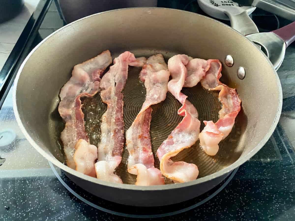 5 Slices of bacon in a frying pan on a glass stove top.