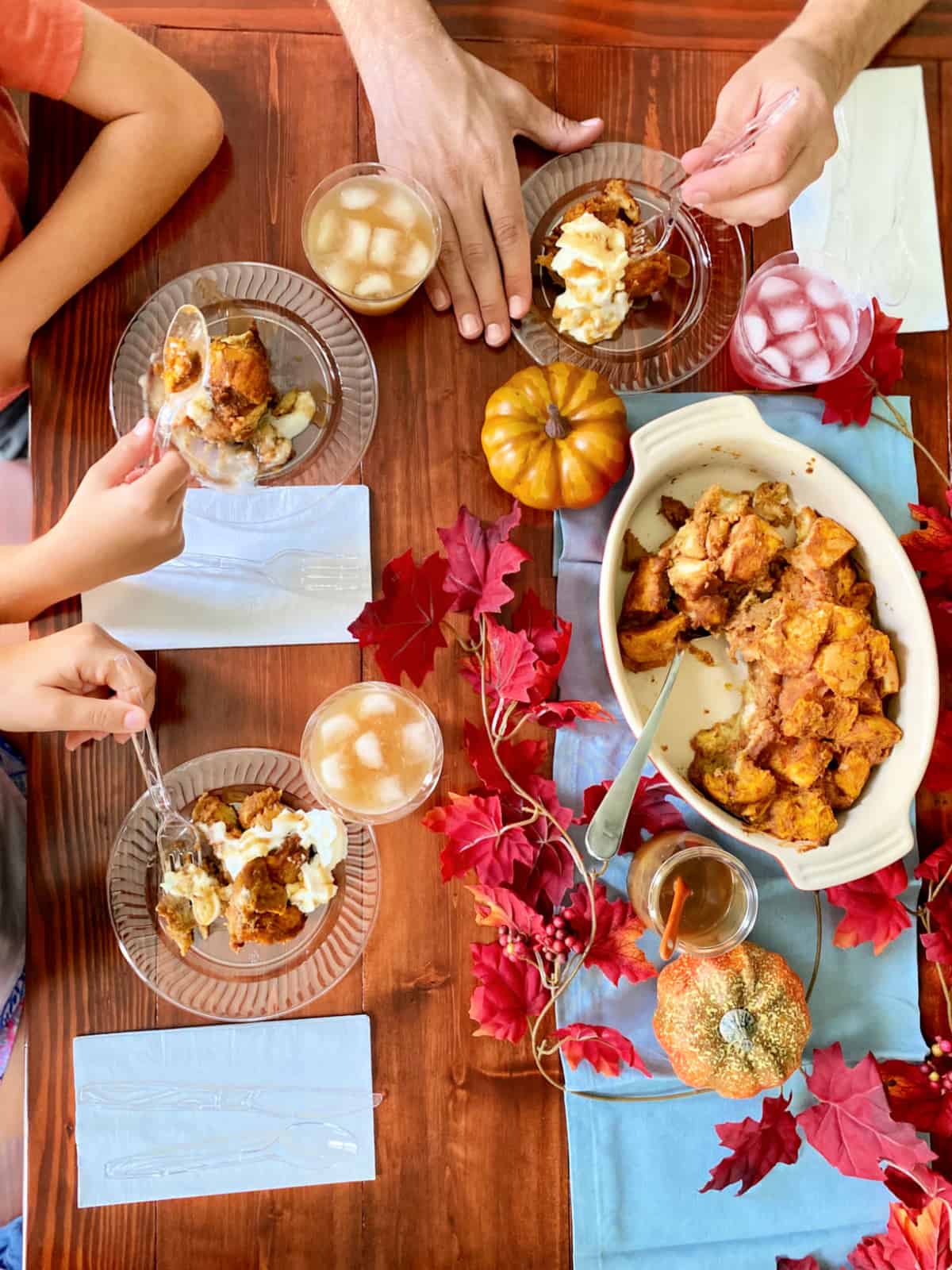 Top view of a bread pudding with fall leaves on a wood table with three plates and hands eating dessert.