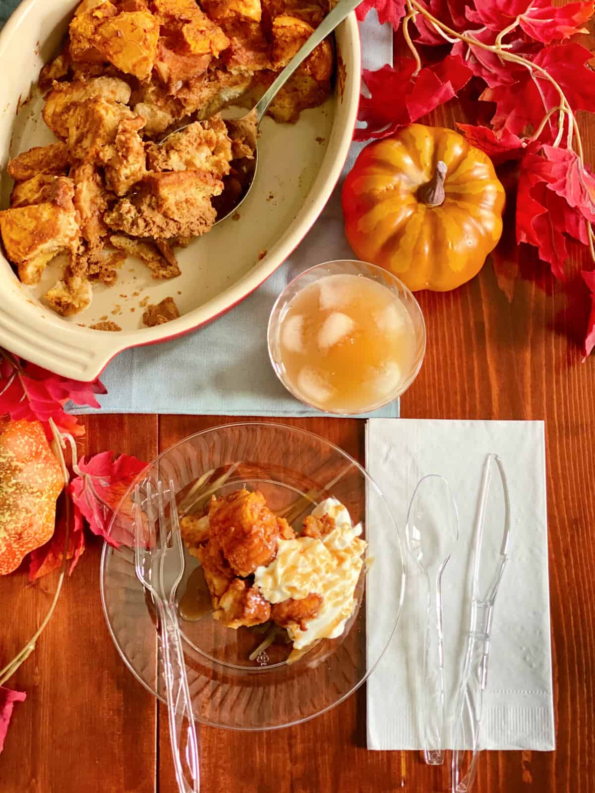 Top view of an oval baking dish with a spoon in it with clear plastic plate, cutlery, and apple cider next to it.