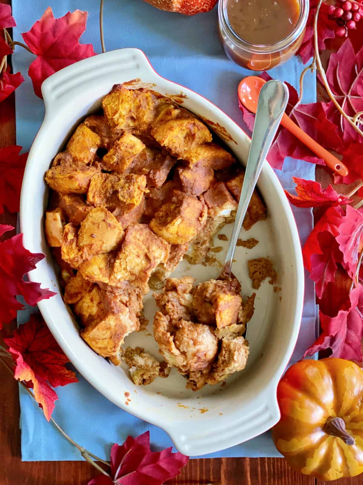 Top view of an oval baking dish with a spoonful of pumpkin bread pudding surrounded by fall leaves and pumpkins.