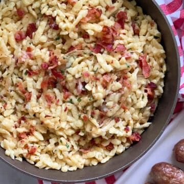 Top view of skillet filled with bacon spaetzle with a red and white striped cloth on the side and brats on the side.