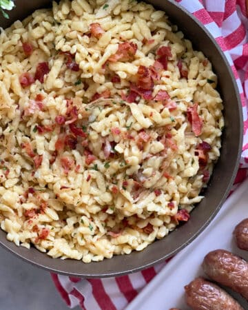 Top view of skillet filled with bacon spaetzle with a red and white striped cloth on the side and brats on the side.