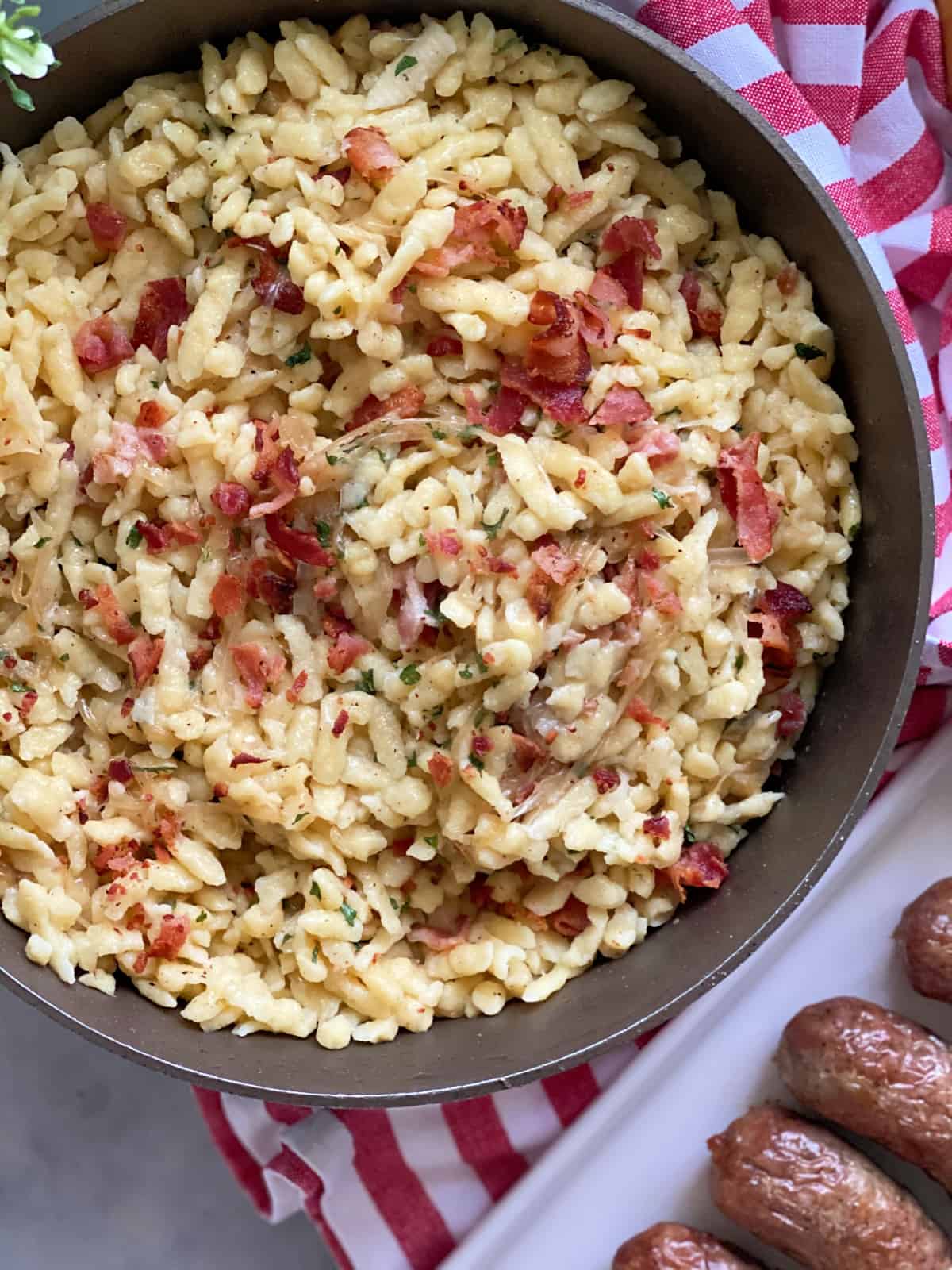 Top view of skillet filled with bacon spaetzle with a red and white striped cloth on the side and brats on the side.