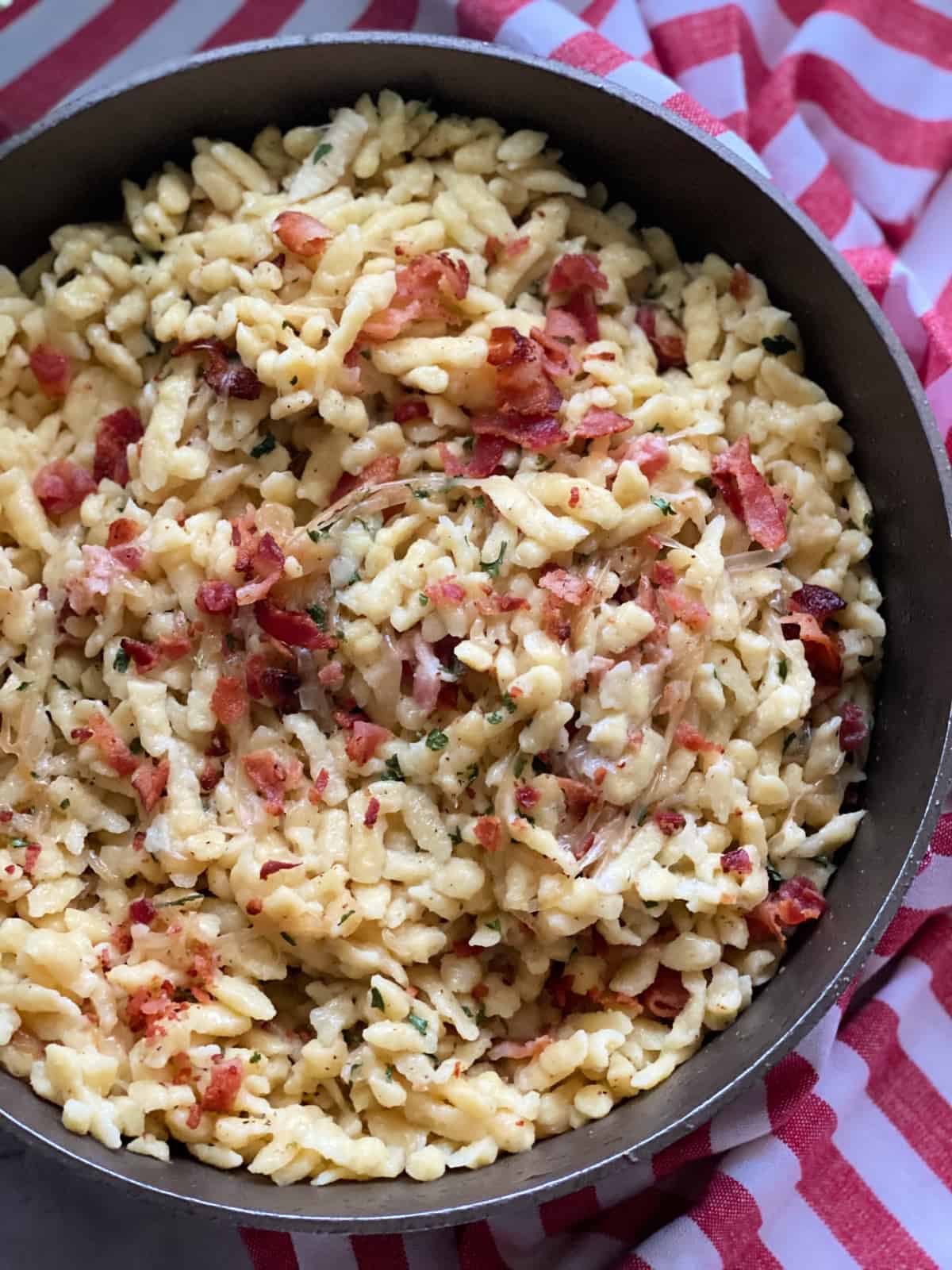 Top view of a skillet filled with bacon spatezle with a red and white cloth wrapped around it.