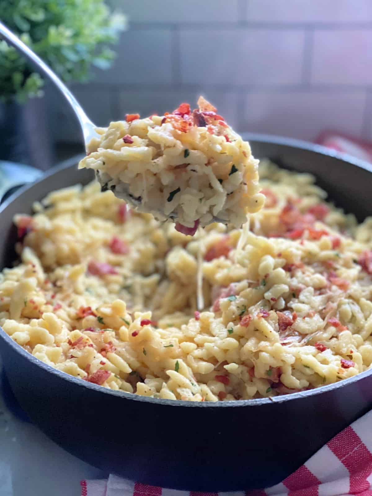 Spoon pulling a scoop of bacon spaetzle from a large skillet filled with spaetzle.