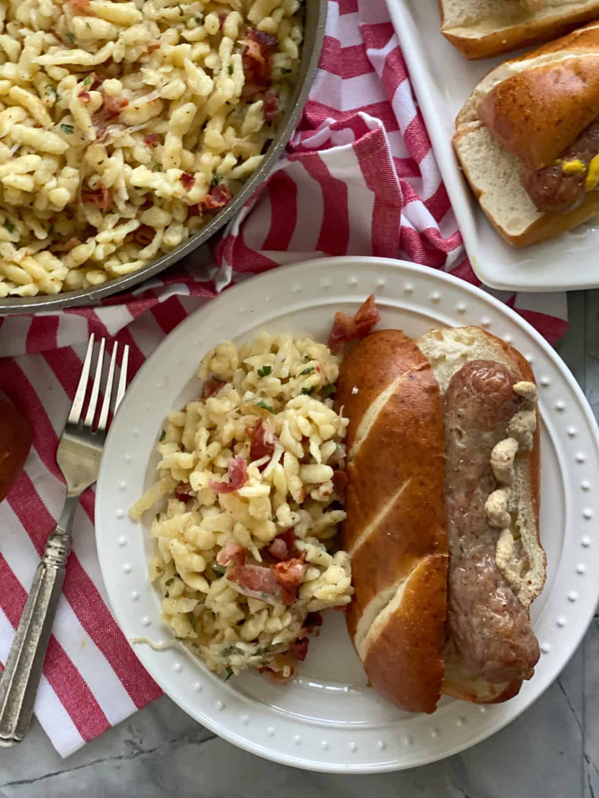 Top view of a white plate filled with spaetzle and a brat on pretzel bun with a red and white striped cloth.