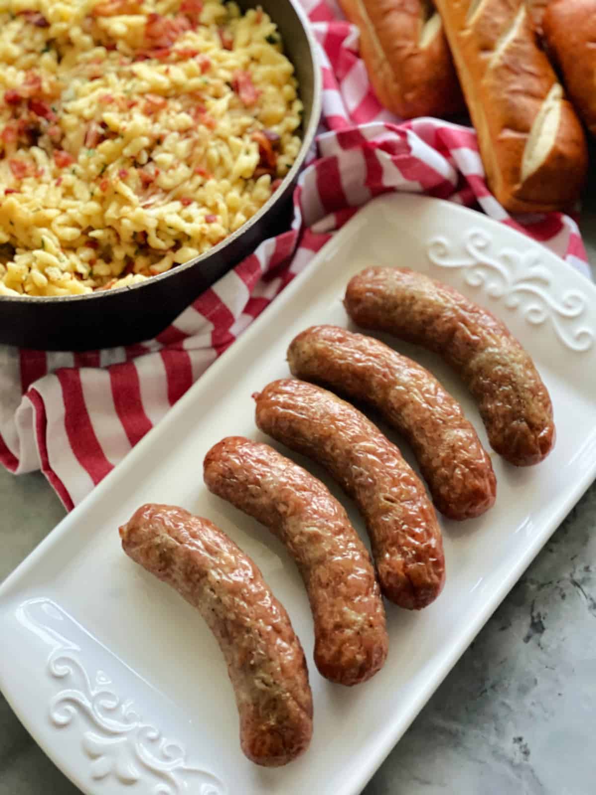 Top view of a white platter filled with five cooked brats and spaetzel on the side.