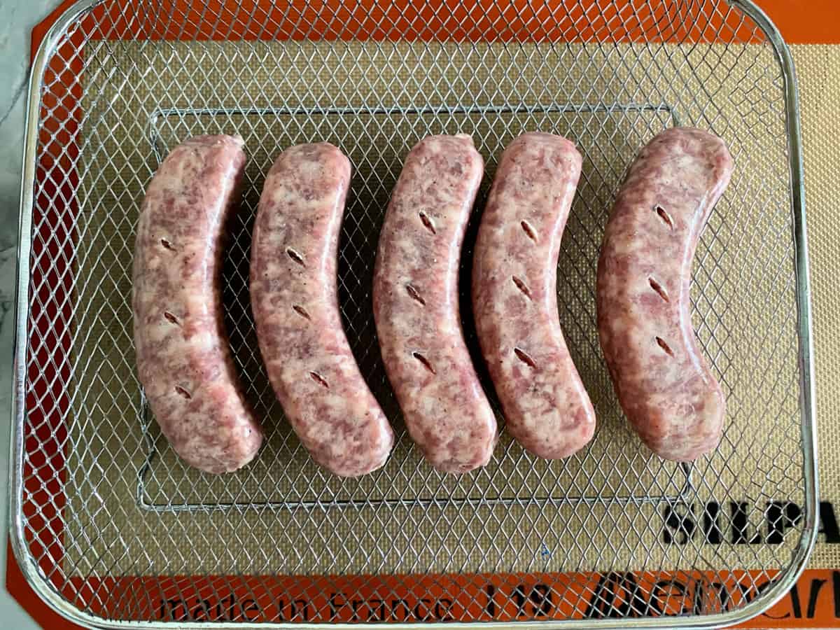 Top view of a wire rack with five raw sausage links with slits in them resting on a silicone mat.