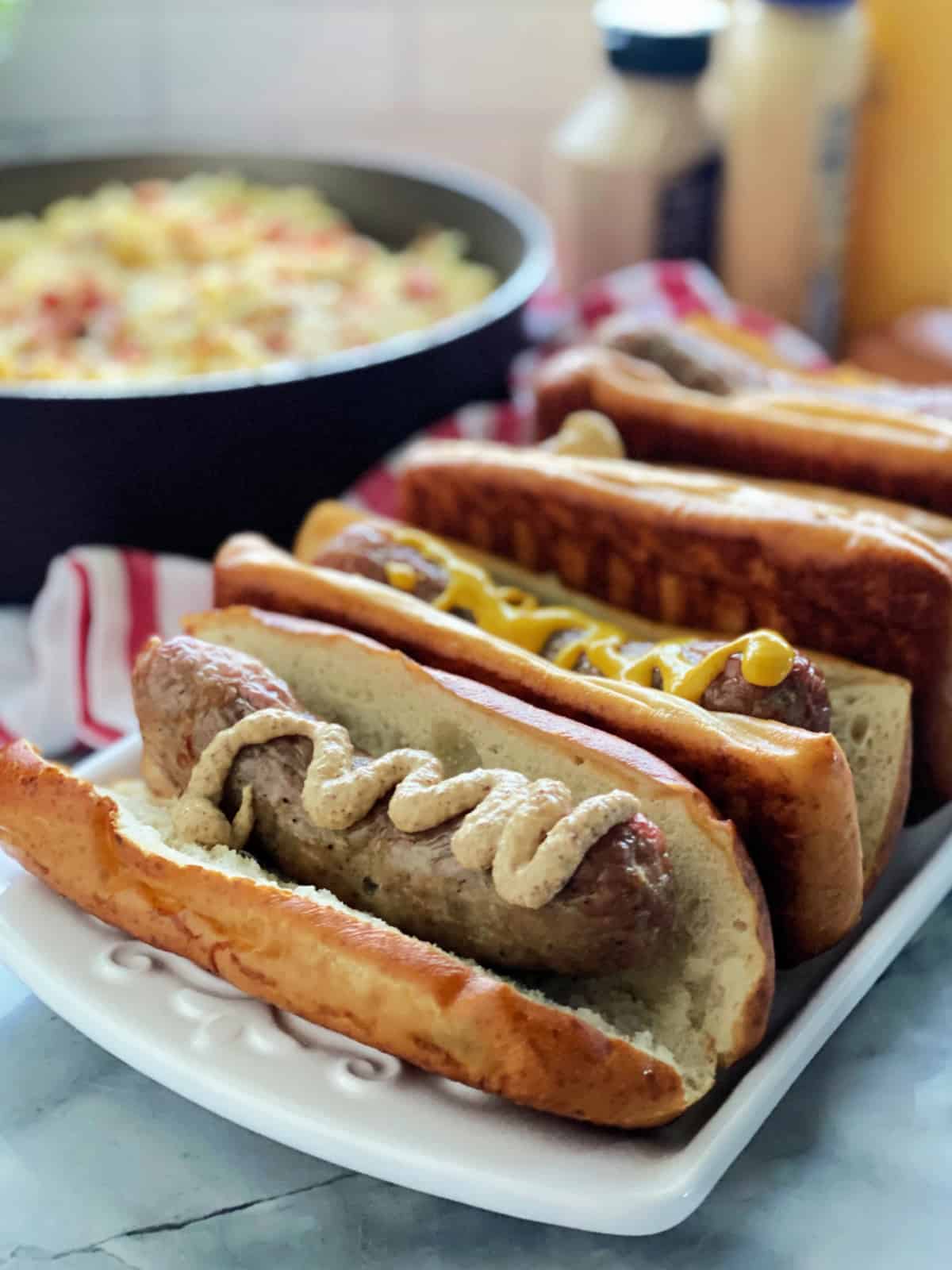 White platter with four pretzel buns filled with brats and mustard.