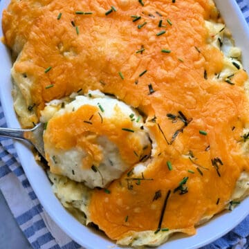 Top view of a white casserole dish filled with mashed potatoes topped with cheddar cheese and chopped chives.