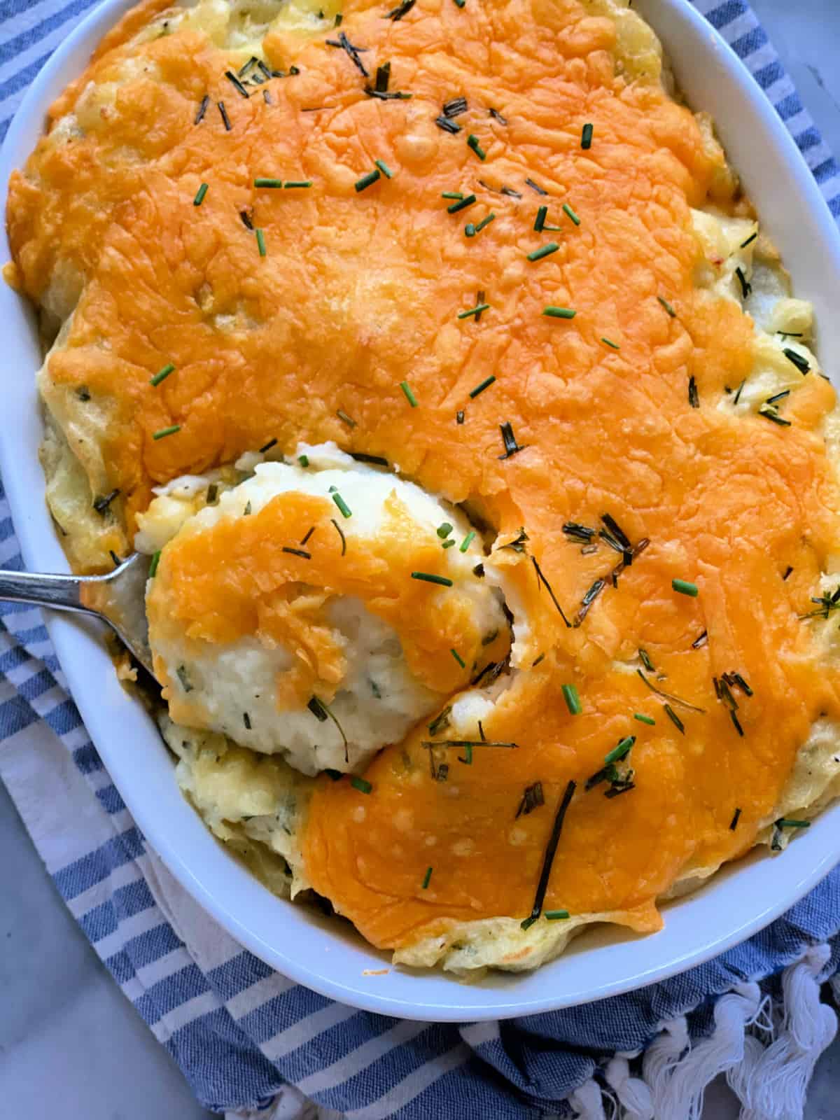 Top view of a white casserole dish filled with mashed potatoes topped with cheddar cheese and chopped chives.