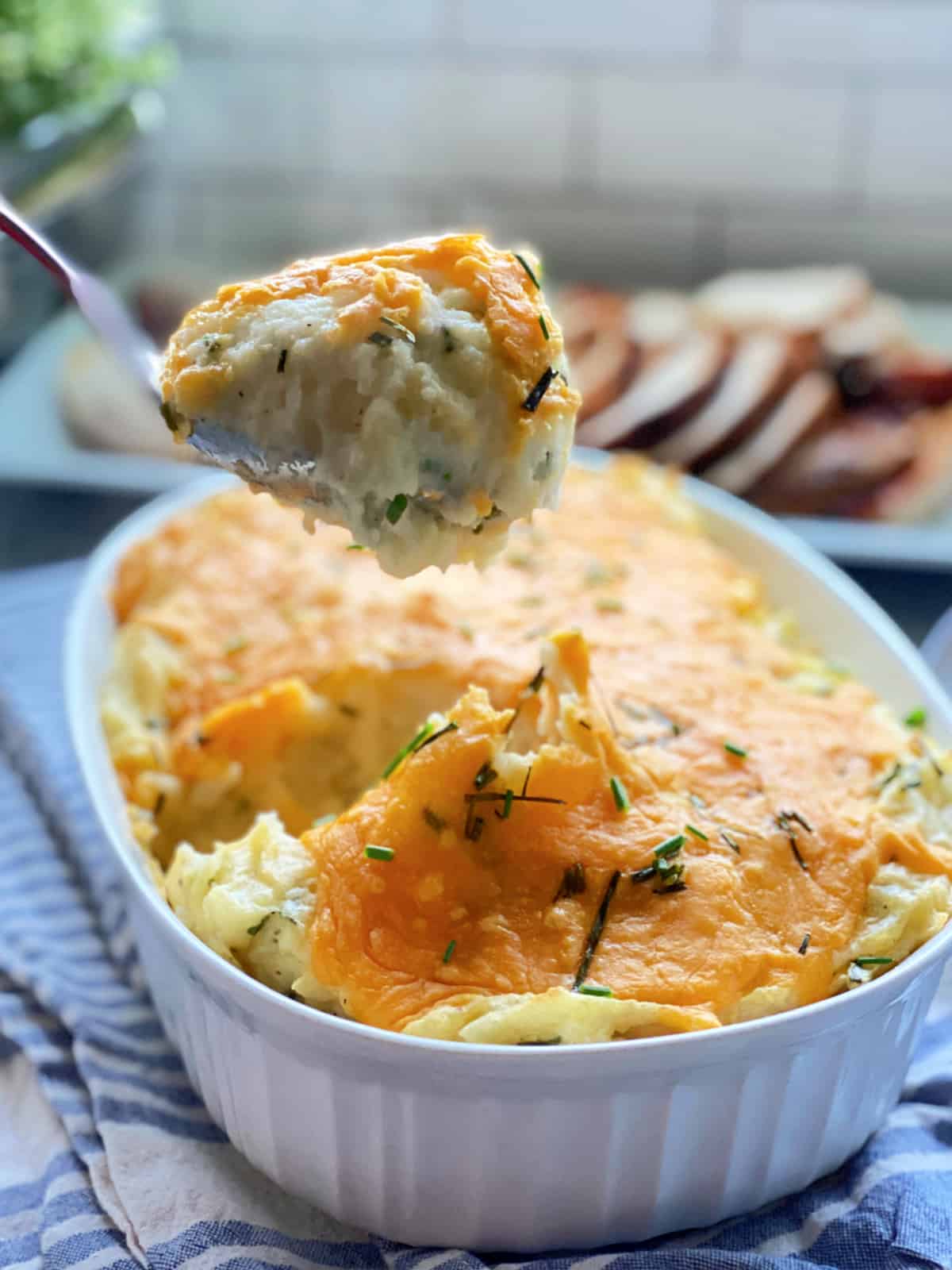 White casserole dish full of Baked Cheddar and Chive Mashed Potatoes with a spoon pulling a scoop of piping hot potatoes from the dish.