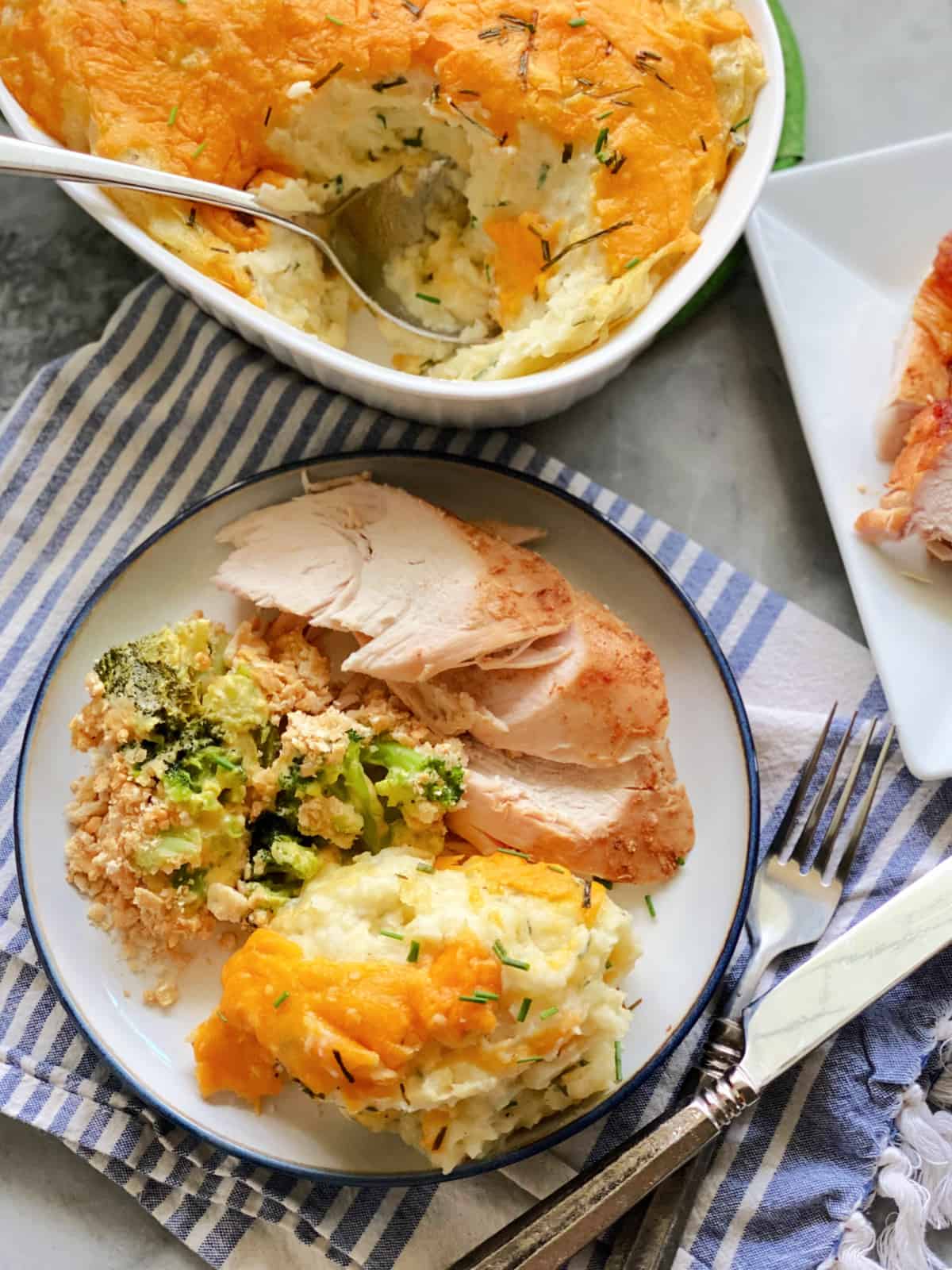 Top view of a white plate filled with mashed potatoes, turkey, broccoli casserole and a casserole dish next to it.