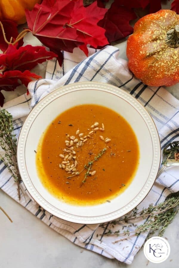 Top view of a white bowl filled with an orange soup with leaves and pumpkins near it with logo on bottom right corner.