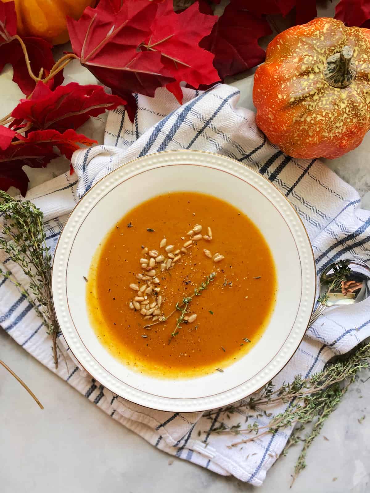 Top view of a white bowl filled with butternut squash soup with pine nuts and fall leaves around it.