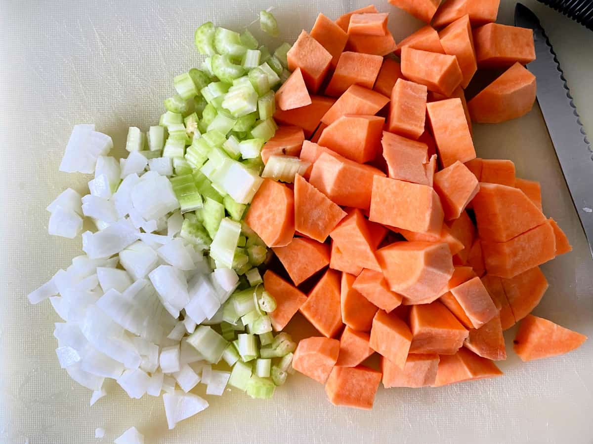 Chopped and peeled sweet potatoes, celery, and onions on a white cutting board with a serated knife next to it.