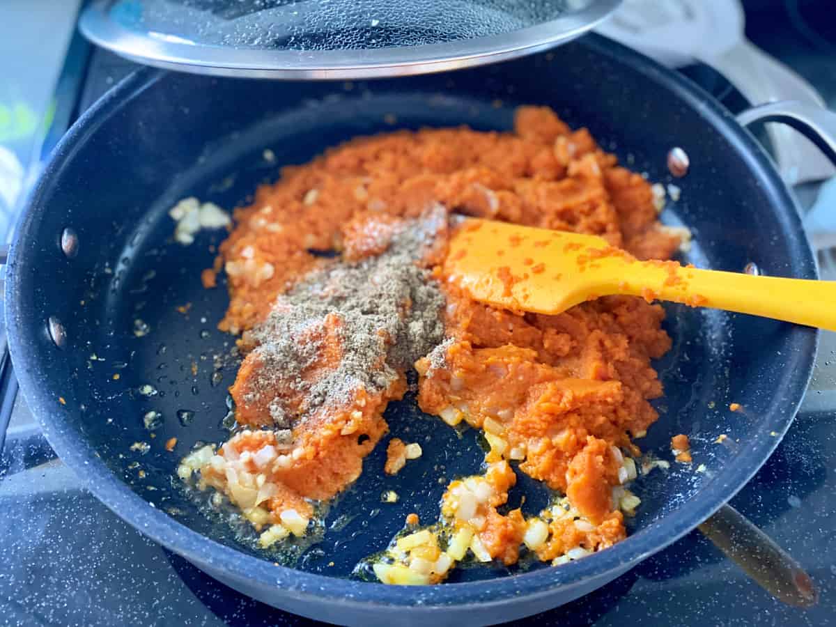 Large black skillet with pumpkin puree, seasonings, onions, and garlic with an orange silicone spatula.