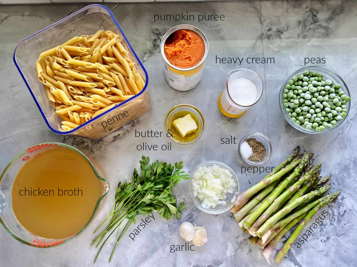 Ingredients on marble counter: penne, chicken broth, parsley, garlic, onion, asparagus, peaas, salt, pepper, pumpkin puree, heavy cream, butter, and olive oil.