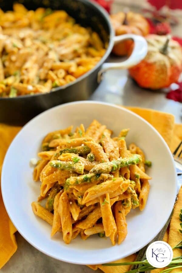 White bowl with creamy pasta with a skillet full of pasta in the background with logo on right bottom corner.