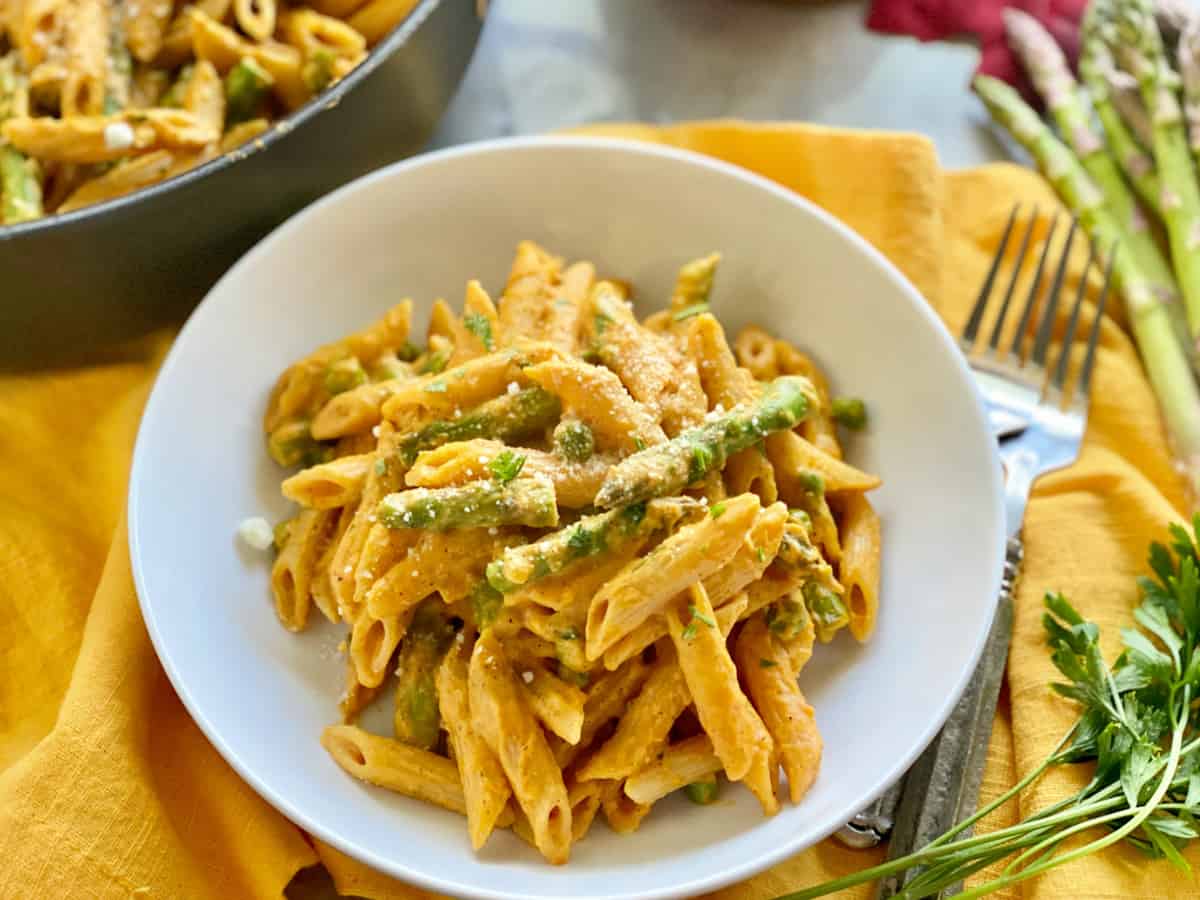 White shallow bowl filled with creamy penne pasta, peas, and asparagus with 2 forks next to the bowl.