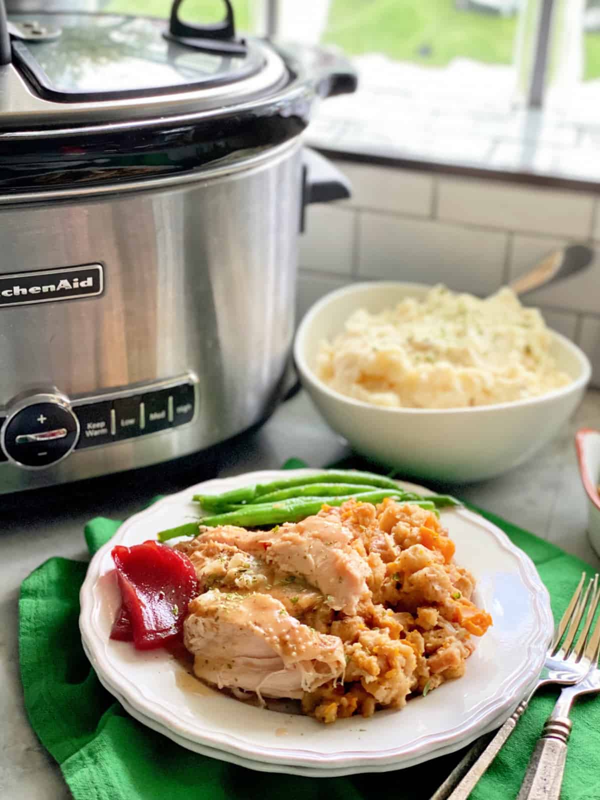 White plate filled with turkey, stuffing, cranberry sauce, and green beans with a slow cooker on the backgournd.