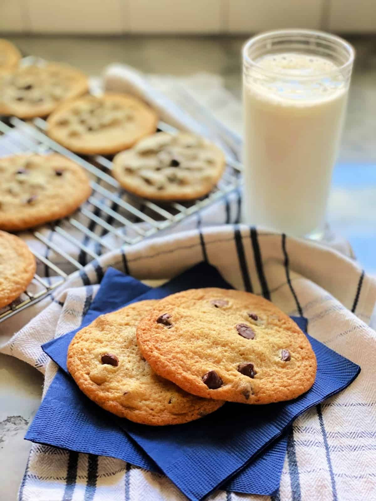Two large thing chocoalte chip cookies stacked on 2 blue napkins with milk in background.