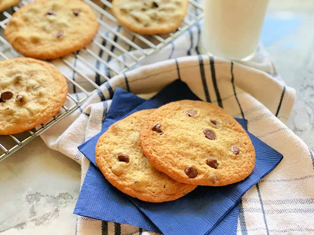 Two dark blue paper napkins with two cookies resting on top of a white and blue dish cloth with milk and cookies in background.