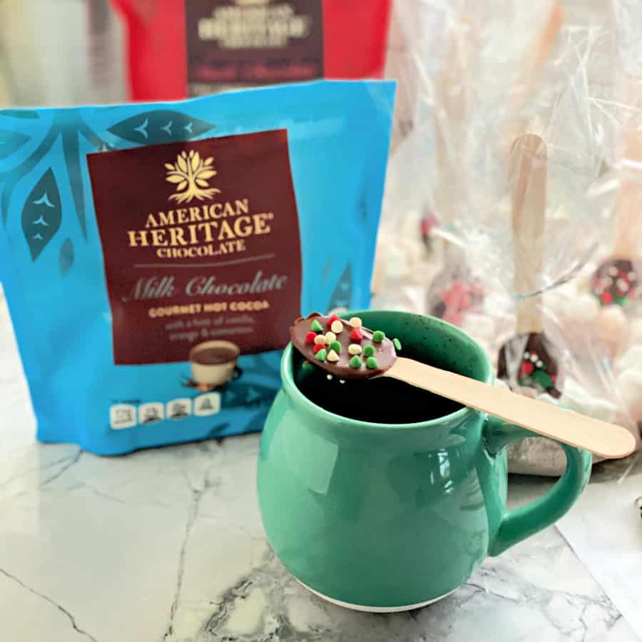 Green mug with a chocolate spoon with sprinkles resting on top with American Heritage chocolate bags in background.