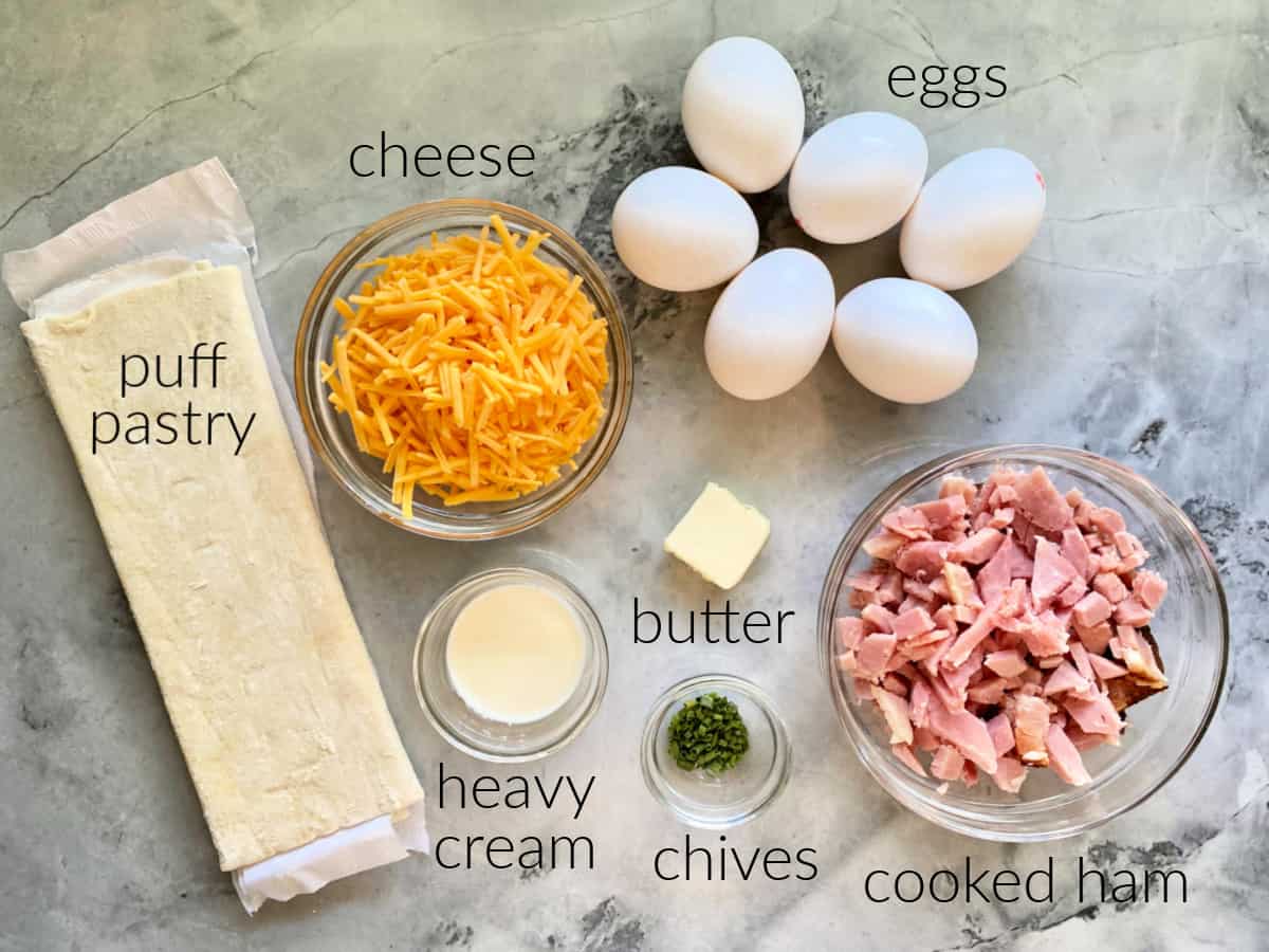 Ingredients on counter: puff pastry, cheese, heavy cream, butter, chives, eggs, and cooked ham.
