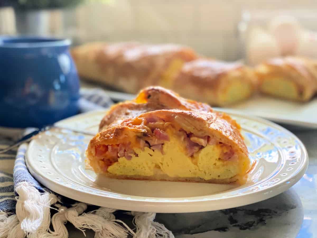 White plate on a blue and white striped cloth napkin with a slice of puff pastry filled with egg, ham, and cheese in it.