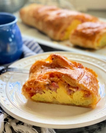 Square picture of a white plate filled with a slice of egg and ham puff pastry with a full pastry in background.