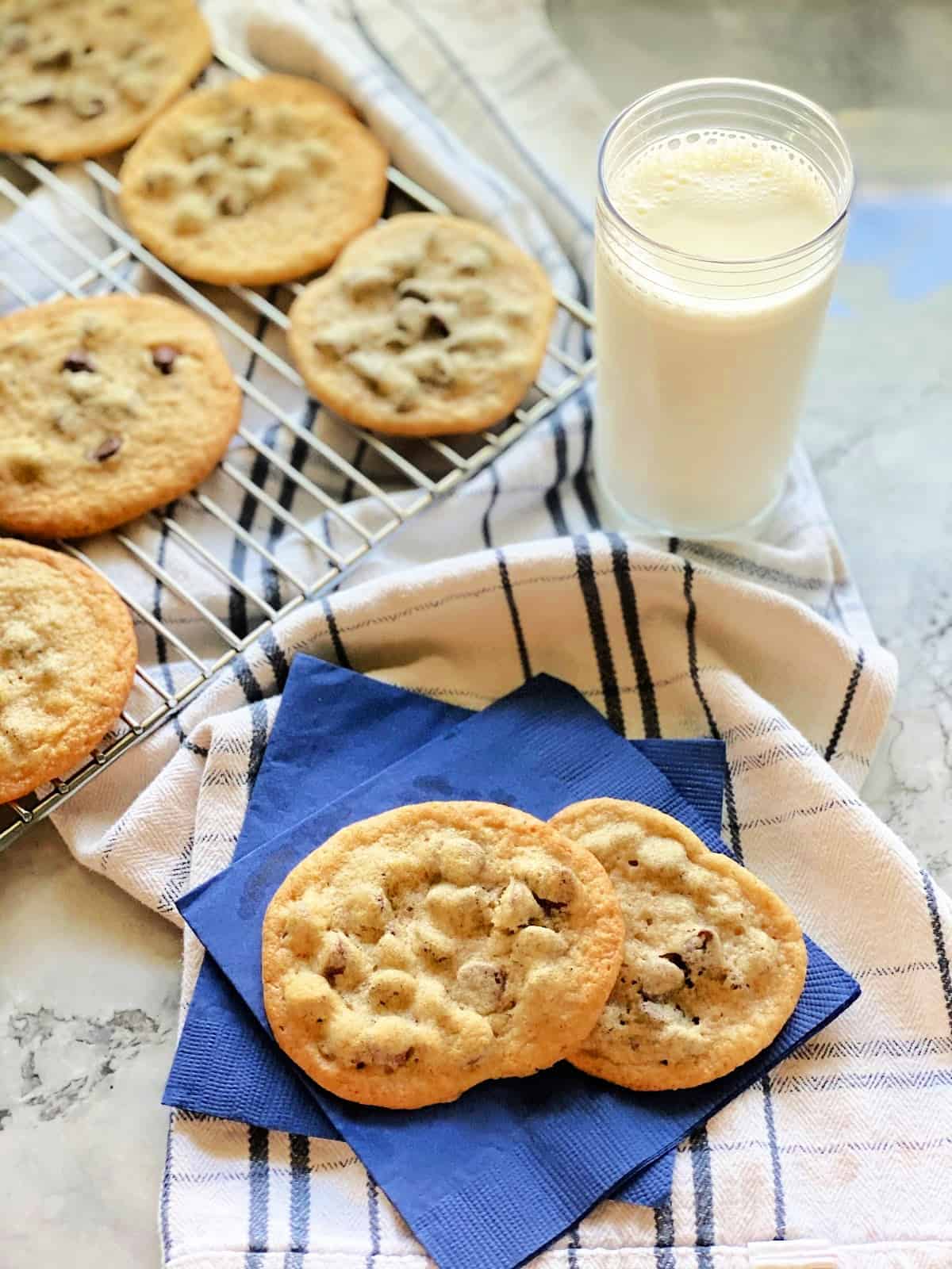 Two blue napkins with two cookies on top with a wire rack of cookies and milk next to it. 